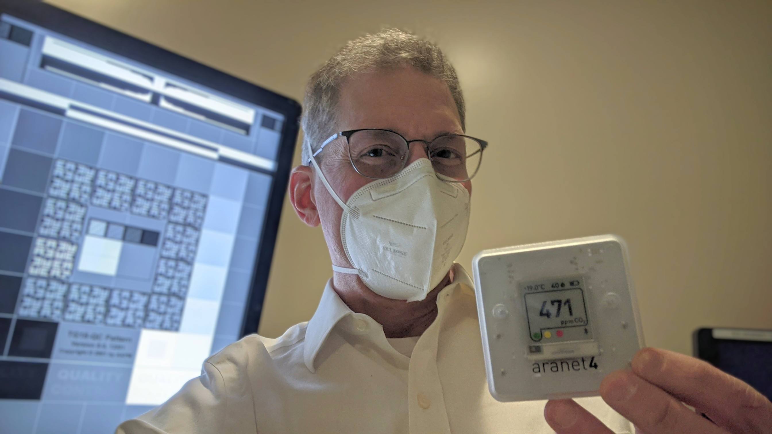 A man with eyeglasses wearing an N95 bifold respirator holds up a CO2 meter displaying 471 ppm. In the background is a radiology monitor with a test patter